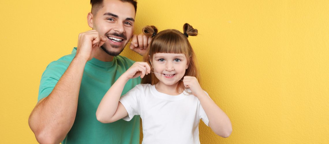 Little girl and her father flossing teeth on color background, space for text